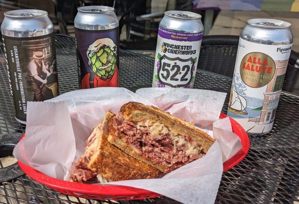 Array of four cans of small batch craft brew beers surround a basket with Reuben sandwich.