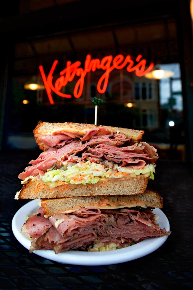 A close up of a Katzinger's reuben with the deli's neon window sign in the background.